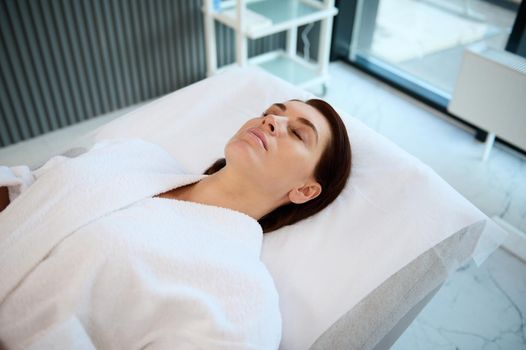 Beautiful Caucasian middle aged woman with perfect glowing skin lying down on a massage table in a spa room, relaxing while receiving a professional skin and body care treatment in wellness spa clinic