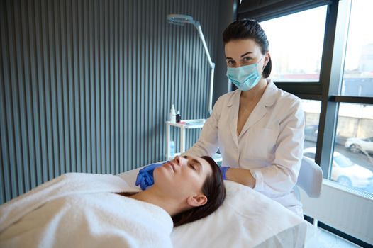 Young cosmetologist performs wellness procedures on the face of a relaxed middle-aged woman lying on a massage couch in a cosmetology spa clinic. Anti-age treatment and professional skin care concept