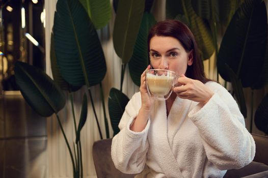 Beautiful European middle-aged woman drinking coffee and talking on mobile phone while resting in luxury wellness spa centre, enjoying beauty procedures. Wellness spa resort, cosmetology concept