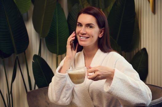 Beautiful woman holds a cup with cappuccino and smiles looking at camera with a beautiful toothy while talking on mobile phone, relaxing in the spa lounge after receiving wellness beauty treatment