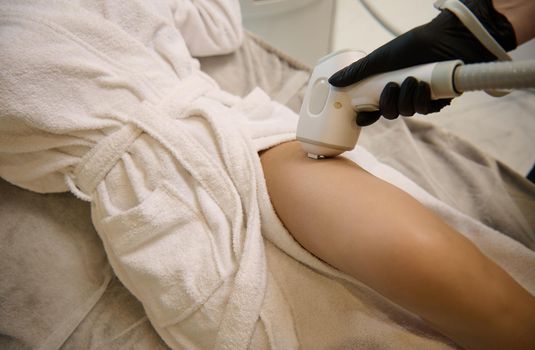 Close-up of a naked leg of woman in white bathrobe receiving hair removal procedure on legs with professional modern laser apparatus in wellness spa clinic. Laser epilation, cosmetology, spa concept