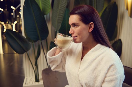 Side portrait of elegant beautiful dark haired middle aged woman in white terry bathrobe drinking cappuccino during coffee break relaxing in spa after professional beauty therapy. Cosmetology concept