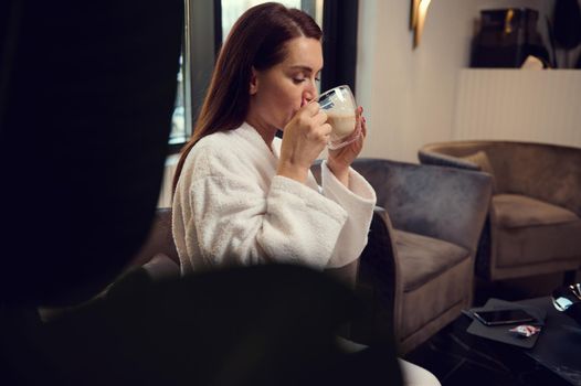 Attractive relaxed European pretty woman enjoying a coffee break while visiting a luxury wellness spa clinic. Beautiful woman drinking cappuccino, sitting on an armchair in a spa salon lounge area