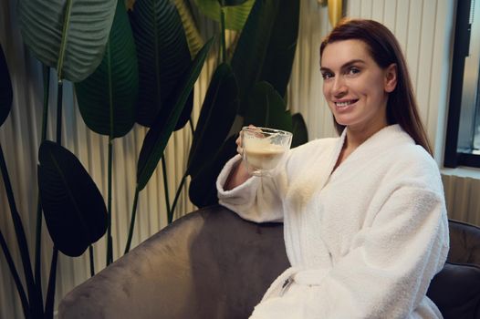 Charming Caucasian middle aged woman in white bathrobe enjoying a coffee break, sitting on an armchair in the lounge of a modern spa clinic, smiles cheerfully looking at camera