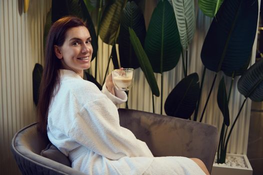 Charming Caucasian middle aged woman in white bathrobe enjoying coffee break, sitting on an armchair in the lounge of a modern spa clinic, smiles cheerfully looking at camera