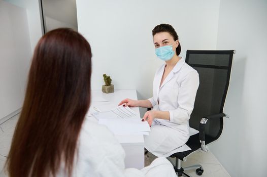 Woman at a doctor's appointment in a white office. Confident general practice cosmetologist beautician wearing medical protective mask reading medical documentation during examination her patient