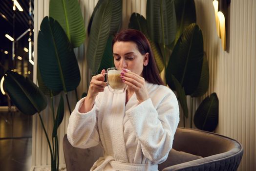 Beautiful woman drinking cappuccino, sitting on an armchair in a spa salon lounge area. Attractive relaxed European pretty woman enjoying a coffee break while visiting a luxury wellness spa clinic.