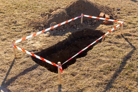 Archaeological excavations in the middle of the field. A small hole in the ground is fenced with red and striped construction tape. Ditch in the ground. Construction works.