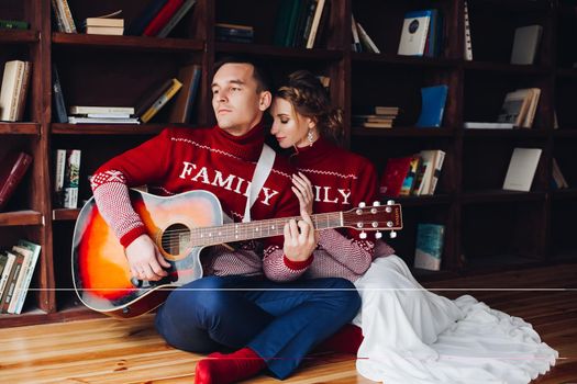 Couple wearing in red sweaters, sitting on floor at library and playing on gitar, and embracing. Happy family romantic spending time together. Winter holidays, x mas, new year concept.