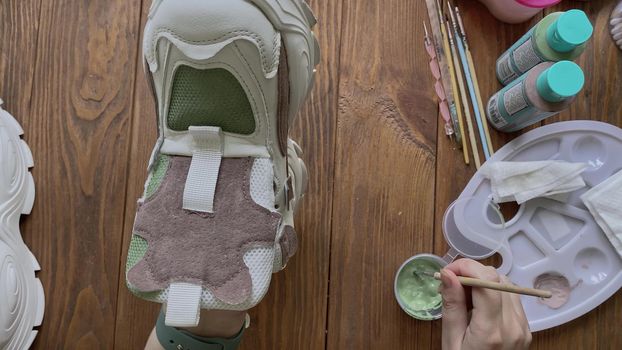 Women's hands paint sneakers with brush on wooden table. Palette with brown and green acrylic on desktop. Designer's workshop. Hobbies, creativity and handmade work at home.