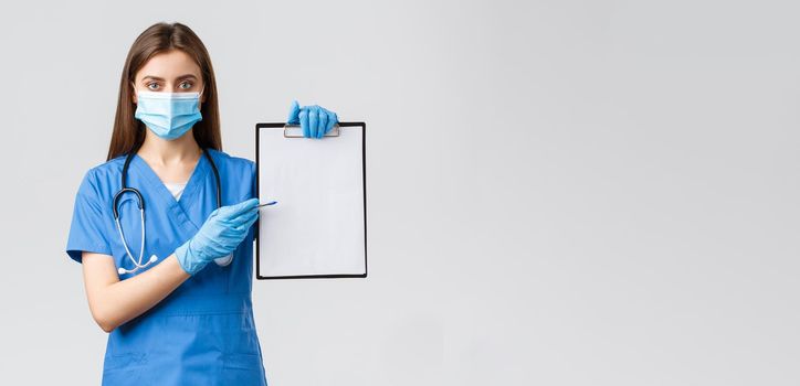 Covid-19, preventing virus, health, healthcare workers and quarantine concept. Serious-looking female nurse or doctor in blue scrubs prescribe medication, pointing at paper on clipboard.