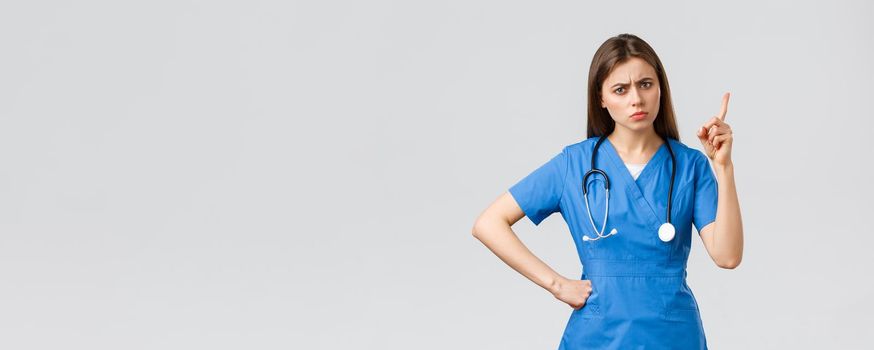 Medical workers, healthcare, covid-19 and vaccination concept. Frowning young female nurse or doctor in blue scrubs, frowning, shaking finger as scolding patient bad behaviour, grey background.