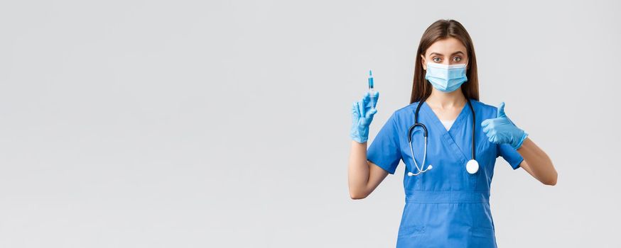 Covid-19, preventing virus, healthcare workers and quarantine concept. Confident female nurse, doctor in blue scrubs, medical mask and gloves assure in quality of coronavirus vaccine, hold syringe.