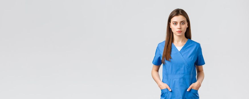 Healthcare workers, prevent virus, insurance and medicine concept. Serious-looking, confident young medical worker, nurse or doctor in blue scrubs hold hands in pockets and looking camera.