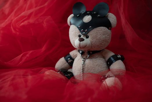toy teddy bear dressed in leather belts and a mask, an accessory for BDSM games against a beautiful red fabric