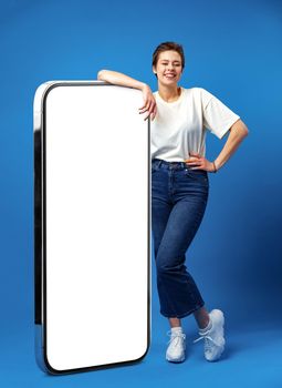 Happy young woman leaning on huge cellphone with empty white screen, for your ad, mock up, blue backgorund