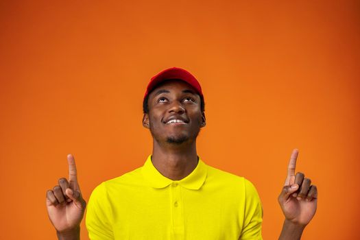 Delivery man in yellow t-shirt and red cap smiling and pointing finger up against orange background, close up