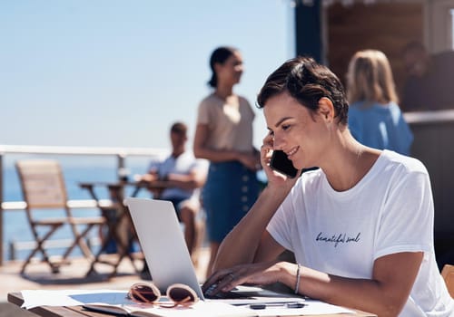 Cropped shot of a cheerful young woman talking on her cellphone while doing work on her laptop next to a beach promenade outside during the day.