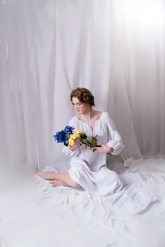 Caucasian Ukrainian woman sitting in the studio in a dress and yellow-blue flowers on a white background during the war. Concept - Stop the war in Ukraine.