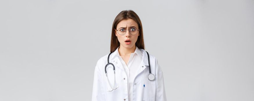 Healthcare workers, medicine, insurance and covid-19 pandemic concept. Shocked nervous female doctor in white scrubs and glasses, doctor gasping and staring concerned camera.