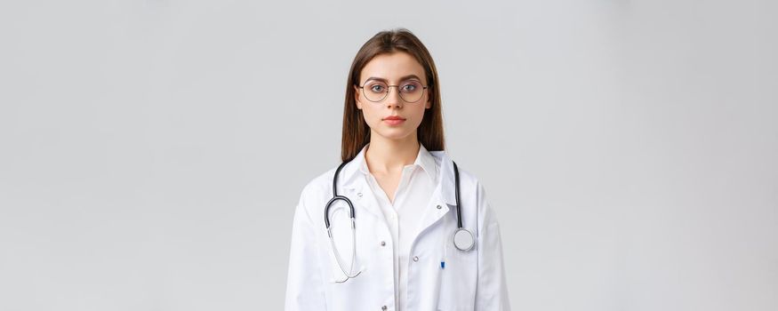 Healthcare workers, medicine, insurance and covid-19 pandemic concept. Serious-looking smart professional doctor, nurse in white scrubs and stethoscope, look camera determined, grey background.