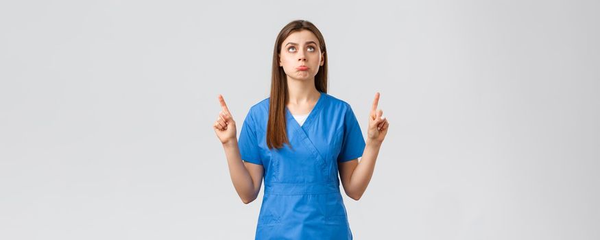 Healthcare workers, prevent virus, covid-19 test screening, medicine concept. Silly upset cute doctor, female nurse in blue scrubs pouting disappointed, pointing and looking up displeased.