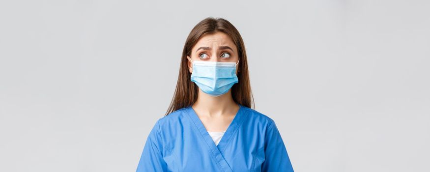 Covid-19, preventing virus, health, healthcare workers and quarantine concept. Skeptical distressed female nurse or doctor in blue scrubs, medical mask, look upper left corner uncertain or puzzled.