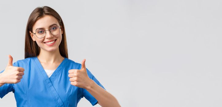 Healthcare workers, medicine, insurance and covid-19 pandemic concept. Excited happy female nurse in blue scrubs, doctor show thumbs-up in support or approval, encourage make tests in clinic.