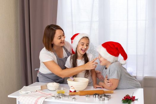 mom and children in light clothes and red hats bake Christmas cookies in the kitchen, on a white table, against the background of a window with white curtains, play merrily with flour