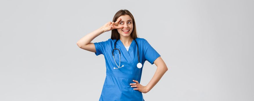 Medical workers, healthcare, covid-19 and vaccination concept. Cheerful and energized young female doctor, pretty nurse in blue scrubs, stethoscope, show peace near eye and smiling upbeat.