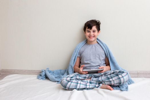 A handsome smiling boy, a teenager in gray pajamas, in a blue knitted plaid sits against a light wall, on the floor, holds a digital tablet, looks at the camera. Copy space