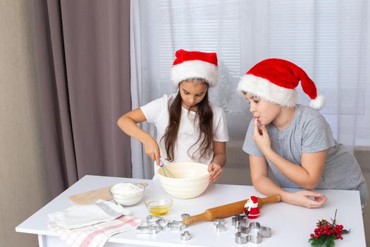two Happy children, brother and sister, in red caps, prepare Christmas cookies in the kitchen. Girl mixes the dough with a whisk in a bowl on a white table