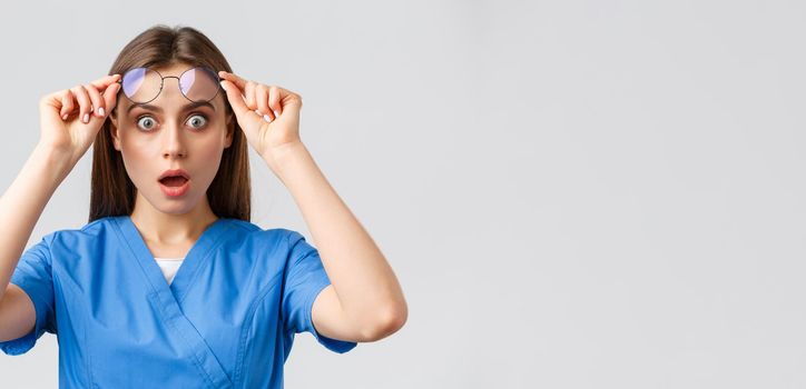 Healthcare workers, medicine, insurance and covid-19 pandemic concept. Surprised or shocked female nurse, doctor in blue scrubs, open mouth and take-off glasses astonished.