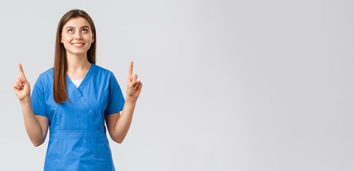 Healthcare workers, prevent virus, covid-19 test screening, medicine concept. Attractive female nurse or doctor in blue scrubs, pointing fingers and looking up with pleased smile, reading banner.