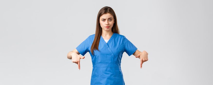 Healthcare workers, prevent virus, covid-19 test screening, medicine concept. Skeptical, disappointed female nurse or doctor in blue scrubs pointing fingers down, smirk unsatisfied, being doubtful.