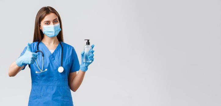 Covid-19, preventing virus, healthcare workers and quarantine concept. Confident female nurse or doctor in blue scrubs, medical mask protective equipment, thumb-up recommend use hand sanitizer.