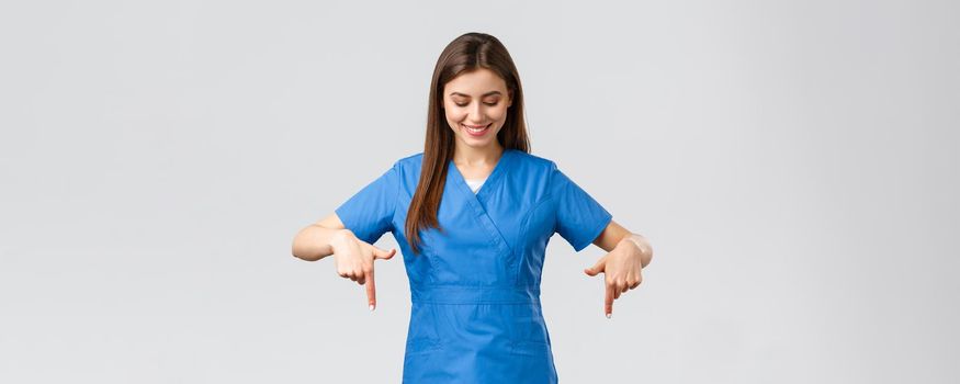 Healthcare workers, prevent virus, covid-19 test screening, medicine concept. Happy and pleased attractive female nurse or doctor, physician in blue scrubs, pointing looking down at advertisement.