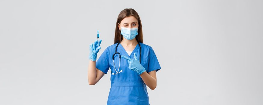 Covid-19, preventing virus, health, healthcare workers and quarantine concept. Cute female nurse or doctor in blue scrubs, medical mask, PPE pointing syringe filled with coronavirus vaccine.