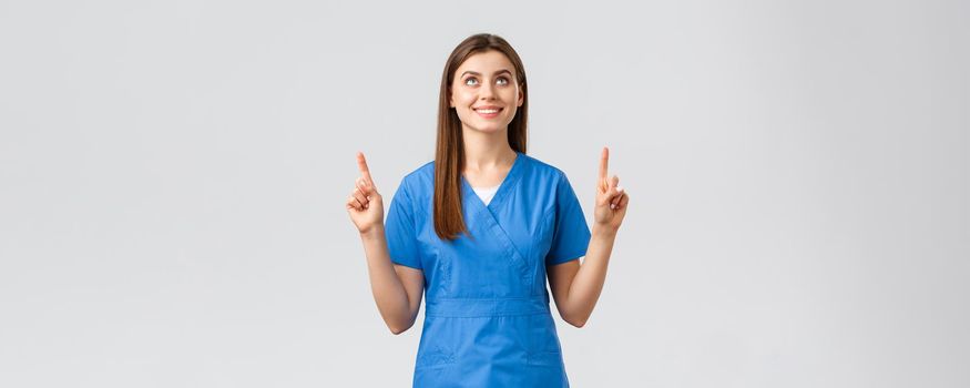 Healthcare workers, prevent virus, covid-19 test screening, medicine concept. Attractive female nurse or doctor in blue scrubs, pointing fingers and looking up with pleased smile, reading banner.