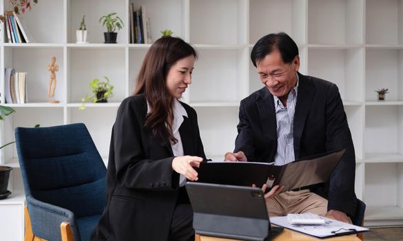 Happy woman and middle-aged man sit at office desk work on laptop together laughing on funny joke, smiling diverse colleagues have fun talking cooperating at workplace.