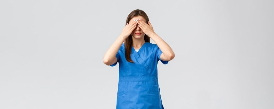 Healthcare workers, prevent virus, insurance and medicine concept. Young woman in blue scrubs, doctor or nurse close eyes with hands, blindfolded medicine, standing grey background.