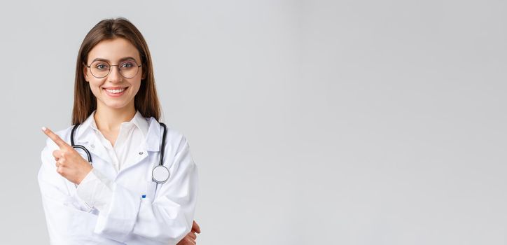 Healthcare workers, medicine and covid-19 pandemic concept. Good-looking professional female doctor in white scrubs and glasses, pointing fingers left, smiling friendly, showing information.