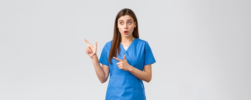 Healthcare workers, prevent virus, insurance and medicine concept. Surprised female nurse or doctor in blue scrubs, looking and pointing fingers upper left corner, gasping amazed.