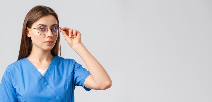Healthcare workers, medicine, insurance and covid-19 pandemic concept. Professional smart female nurse, doctor or intern in blue scrubs and glasses looking at camera determined, grey background.