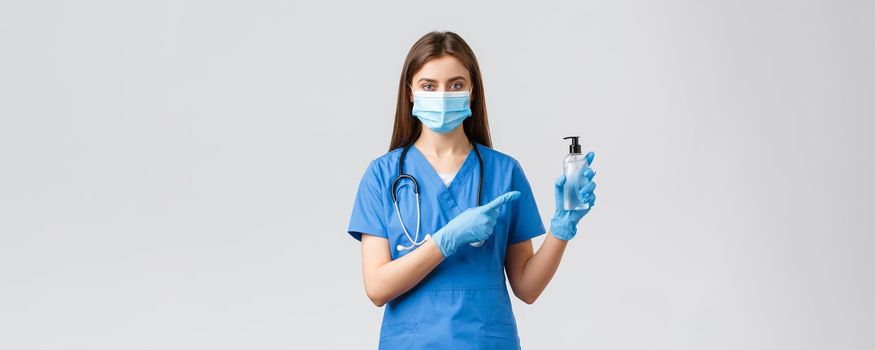 Covid-19, preventing virus, health, healthcare workers and quarantine concept. Young professional female nurse or doctor in blue scrubs, medical mask, pointing finger at hand sanitizer.