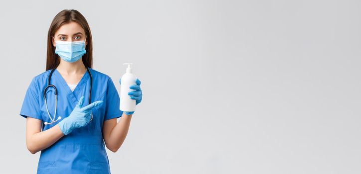 Covid-19, preventing virus, health, healthcare workers and quarantine concept. Young pretty female nurse or doctor in blue scrubs, medical mask and gloves, pointing at hand sanitizer, soap.