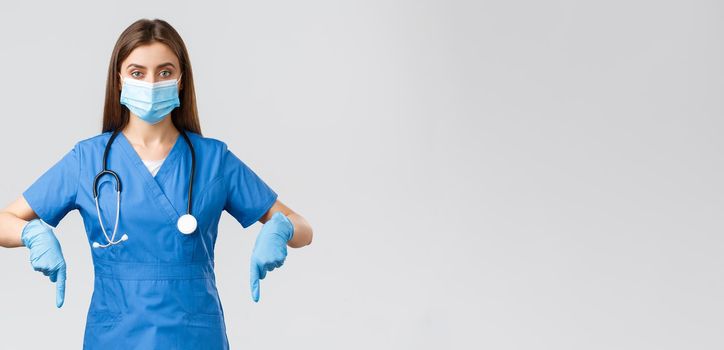 Covid-19, preventing virus, health, healthcare workers and quarantine concept. Determined female nurse or doctor in blue scrubs, medical mask and gloves, pointing fingers down, showing info banner.