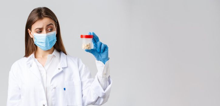 Covid-19, preventing virus, healthcare workers and quarantine concept. Unsure, curious doctor in scrubs and medical mask look intrigued and uncertain at container with medicine pills.