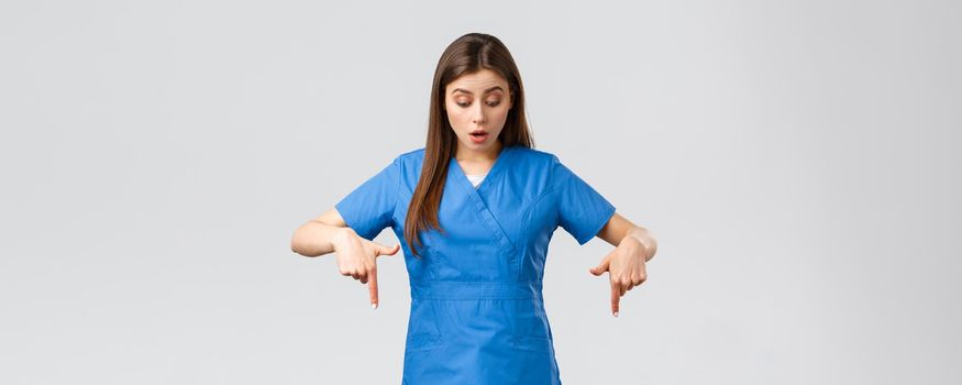 Healthcare workers, prevent virus, covid-19 test screening, medicine concept. Impressed, curious female nurse or doctor in blue scrubs, pointing fingers down and looking at banner wondered.
