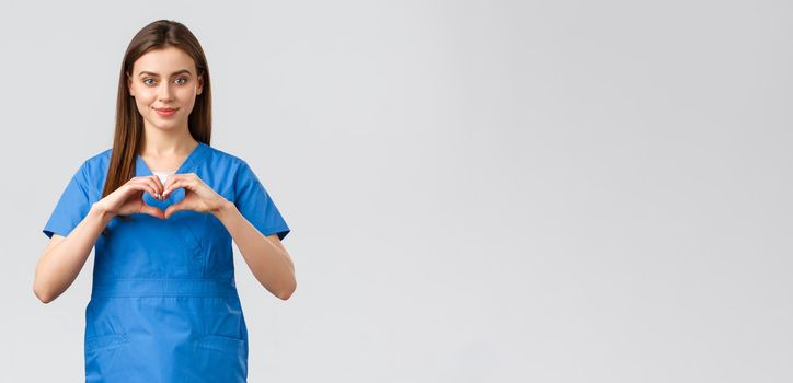 Healthcare workers, prevent virus, insurance and medicine concept. Smiling attractive female doctor, nurse in blue scrubs stay safe home, show heart sign, express respect to coronavirus fighters.